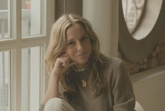 Vrouw met lange, golvende haren, zittend in een lichte ruimte met een ontspannen uitstraling.