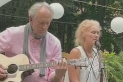 Een man speelt gitaar terwijl een vrouw zingt, beide in een groene omgeving.